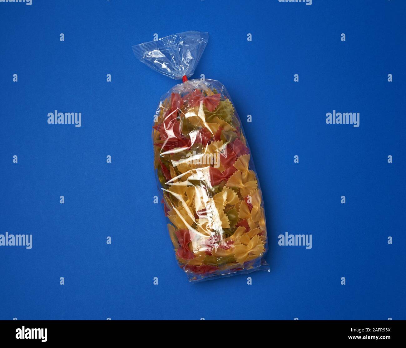 transparent cellophane bag with raw colorful pasta in the form of bows on a blue surface, copy space Stock Photo