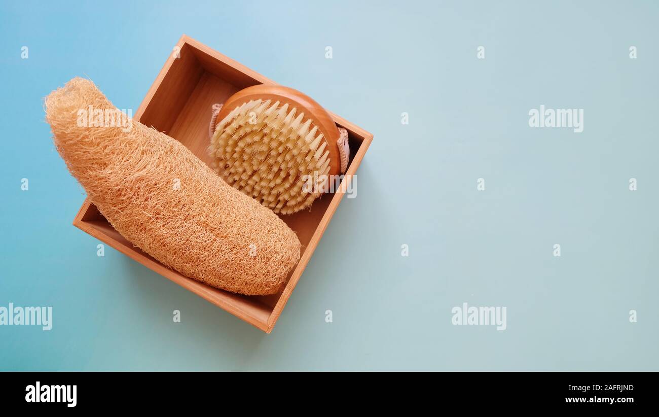 Top view of a round bath brush and a luffa sponge inside a wooden box, on a blue background. Copy space available on the right. Stock Photo