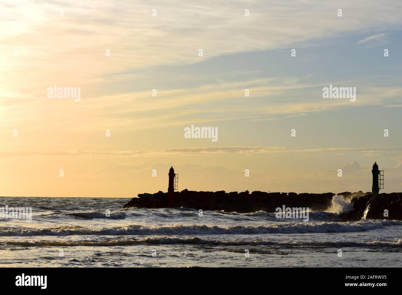Dramatic sunset by the sea with a red sky reflecting on the sea and the waves behind the silhouette of two small lighthouses on the coast Stock Photo