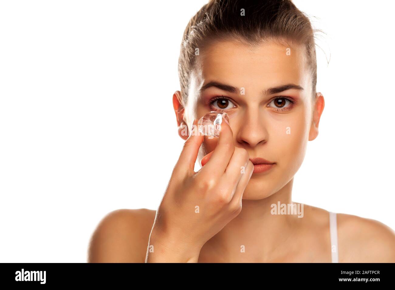 Portrait of young beautiful holding ice cube under her eye on white