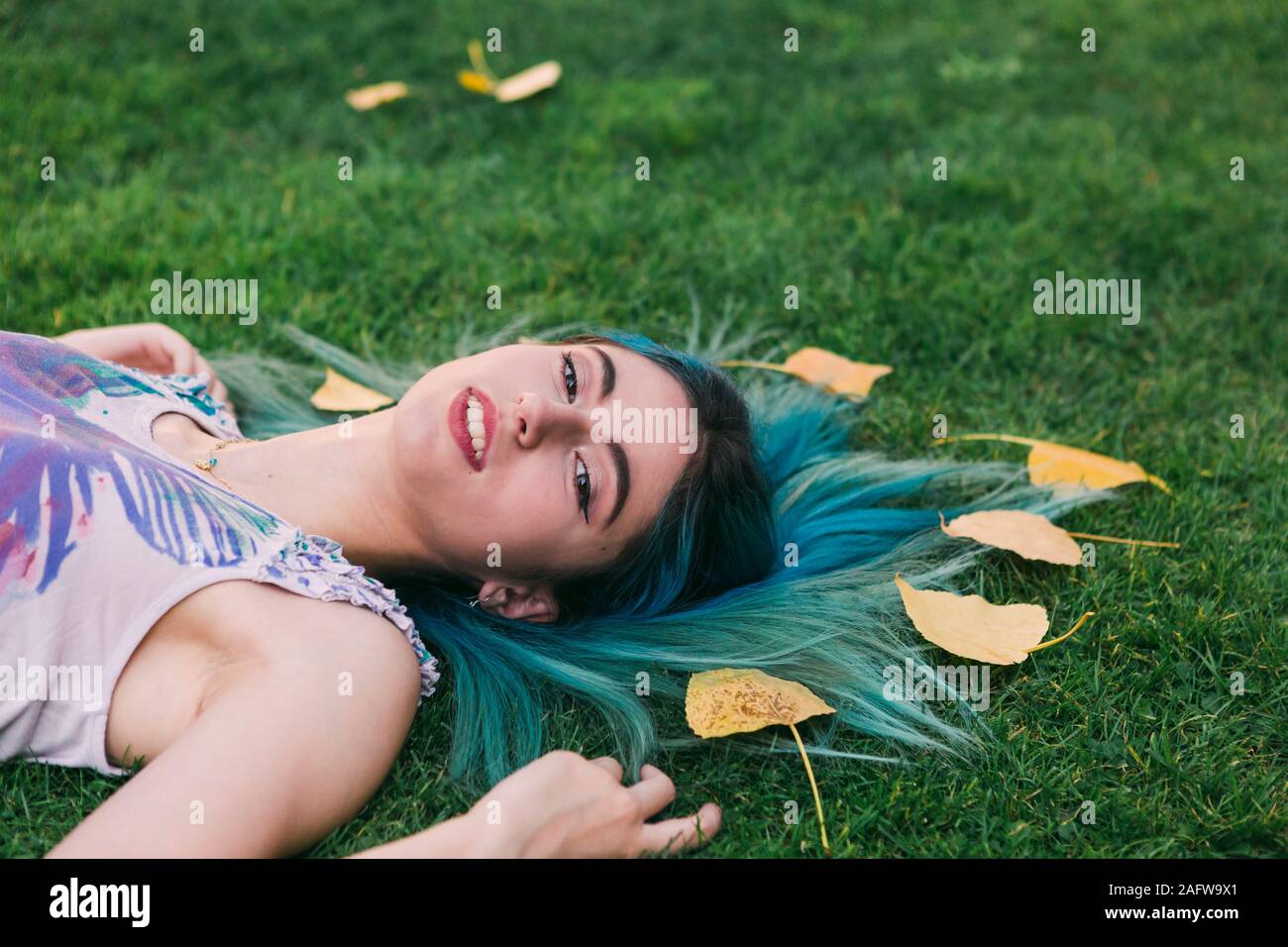 Portrait serene young woman with blue hair laying in grass with autumn leaves Stock Photo