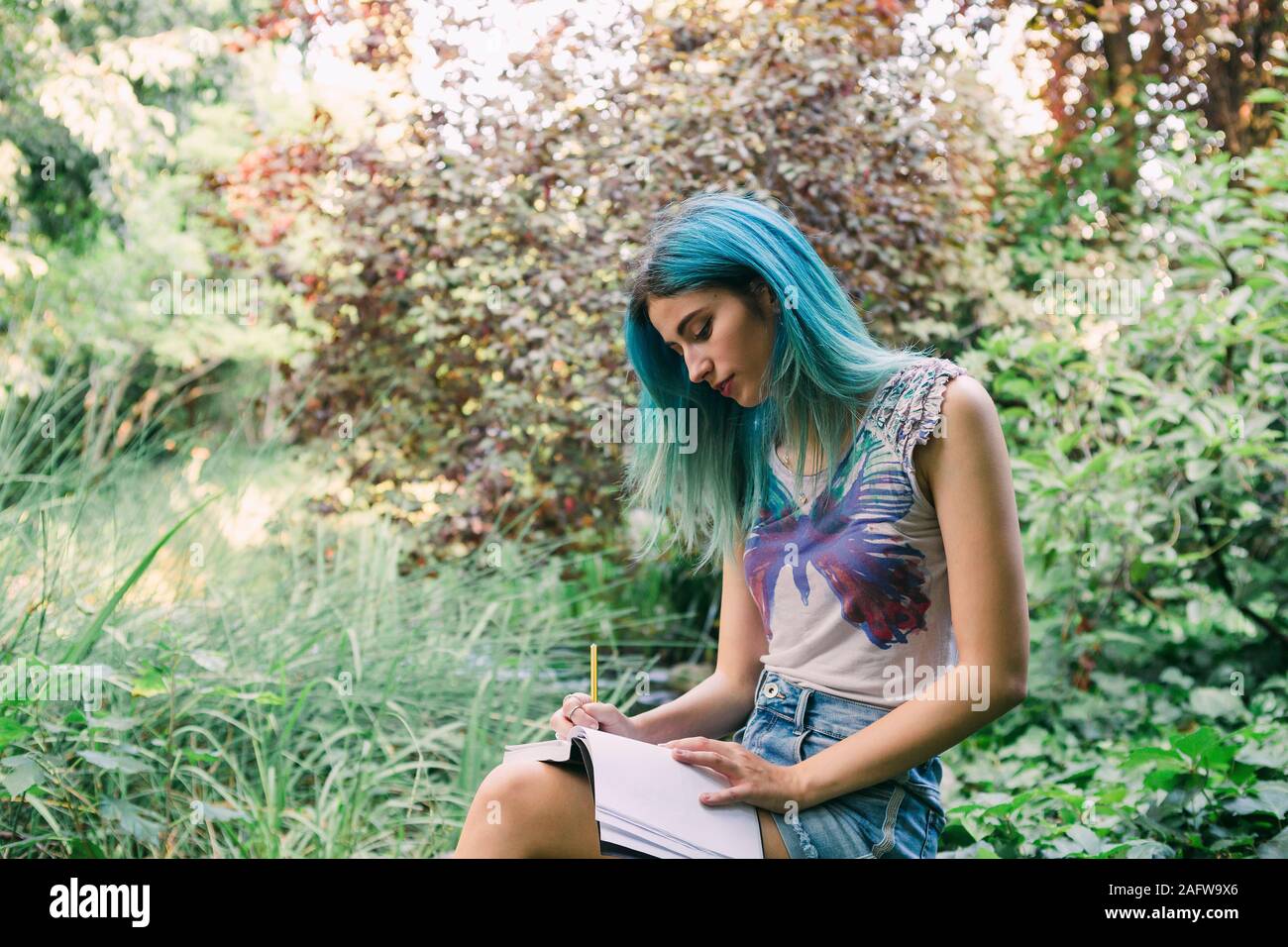 Young woman with blue hair writing in journal in park Stock Photo