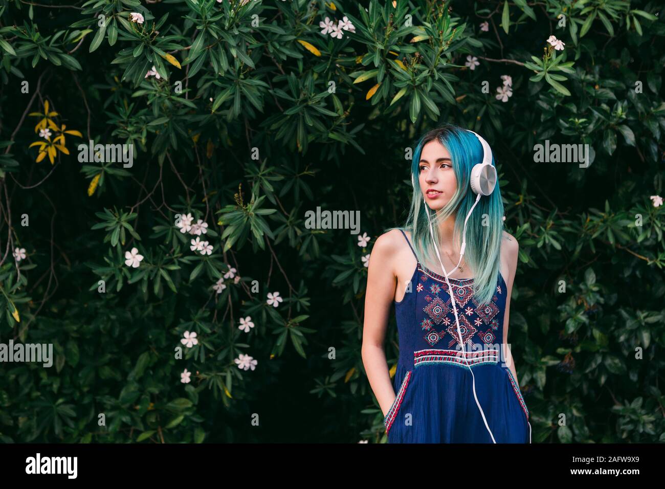 Young woman with blue hair listening to music with headphones in front of flowering tree Stock Photo