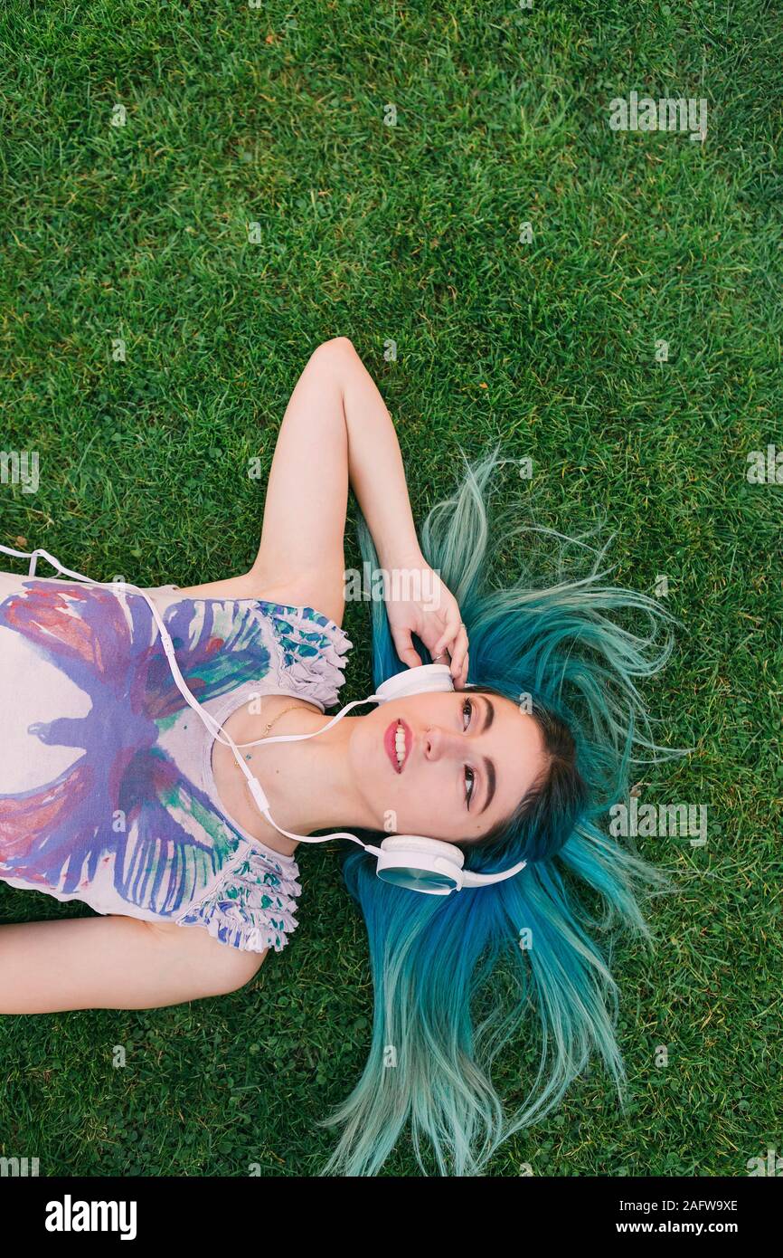 Overhead portrait young woman with blue hair listening to music with headphones, laying in grass Stock Photo