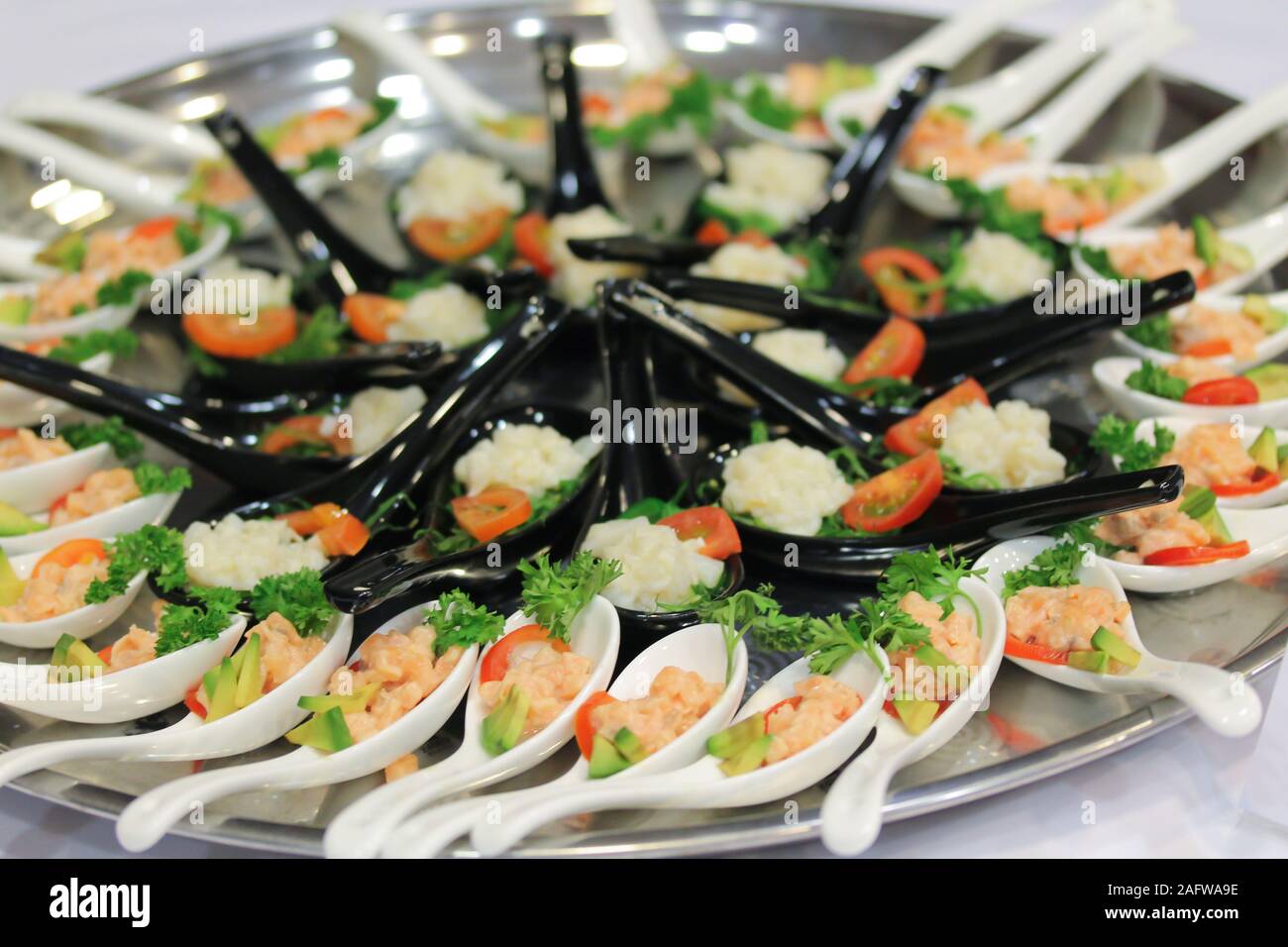Salad snacks in ceramic black and white spoons at the stand-up party or buffet lunch. Tasty food treats with fresh vegetables on big round metal or silver plate onthe table. Catering service Stock Photo