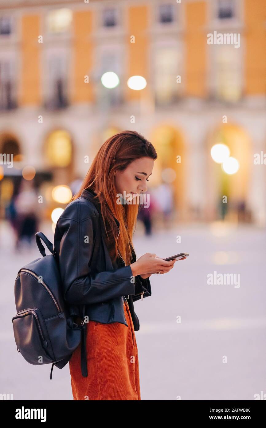 Young woman with backpack using smart phone on urban sidewalk Stock Photo
