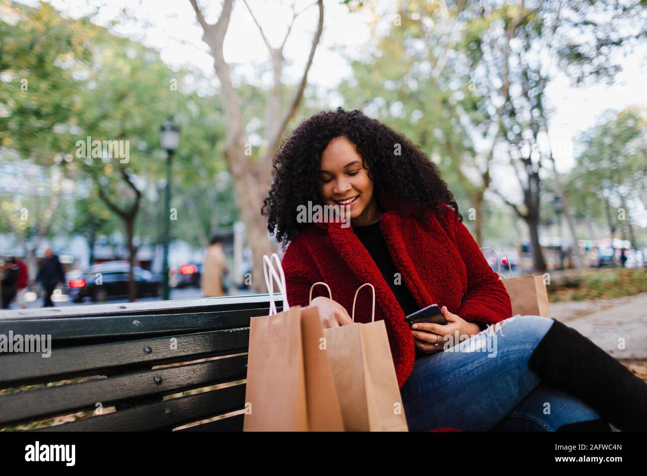 Smiling young woman looking in shopping bags on park bench Stock Photo