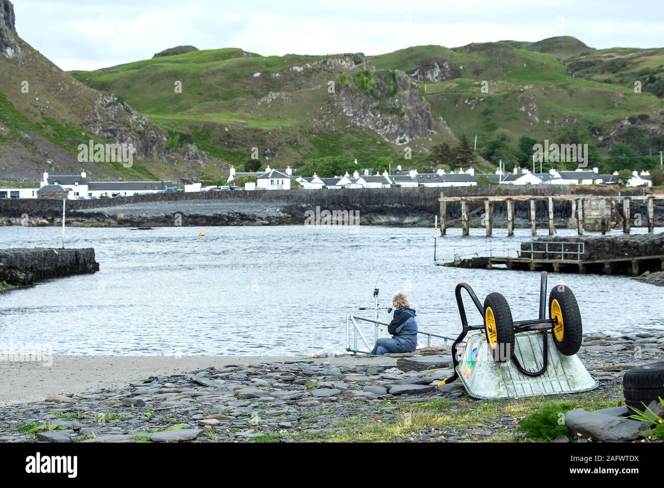 A womand looking back towards the harbour of Ellenabeich, a small village on the isle of Seil  from Easdale Island.  The wheelbarrow in the foreground Stock Photo