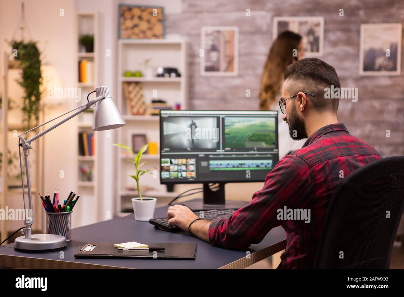 Back view of creative filmmaker working on a movie on laptop. Girlfriend in the background. Stock Photo