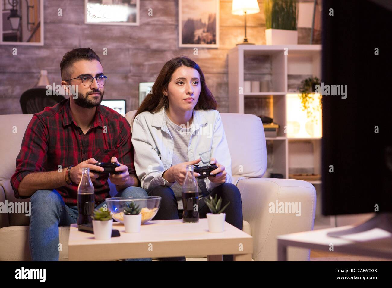 Beautiful young couple having fun playing video games with controllers at night. Couple sitting on sofa. Stock Photo