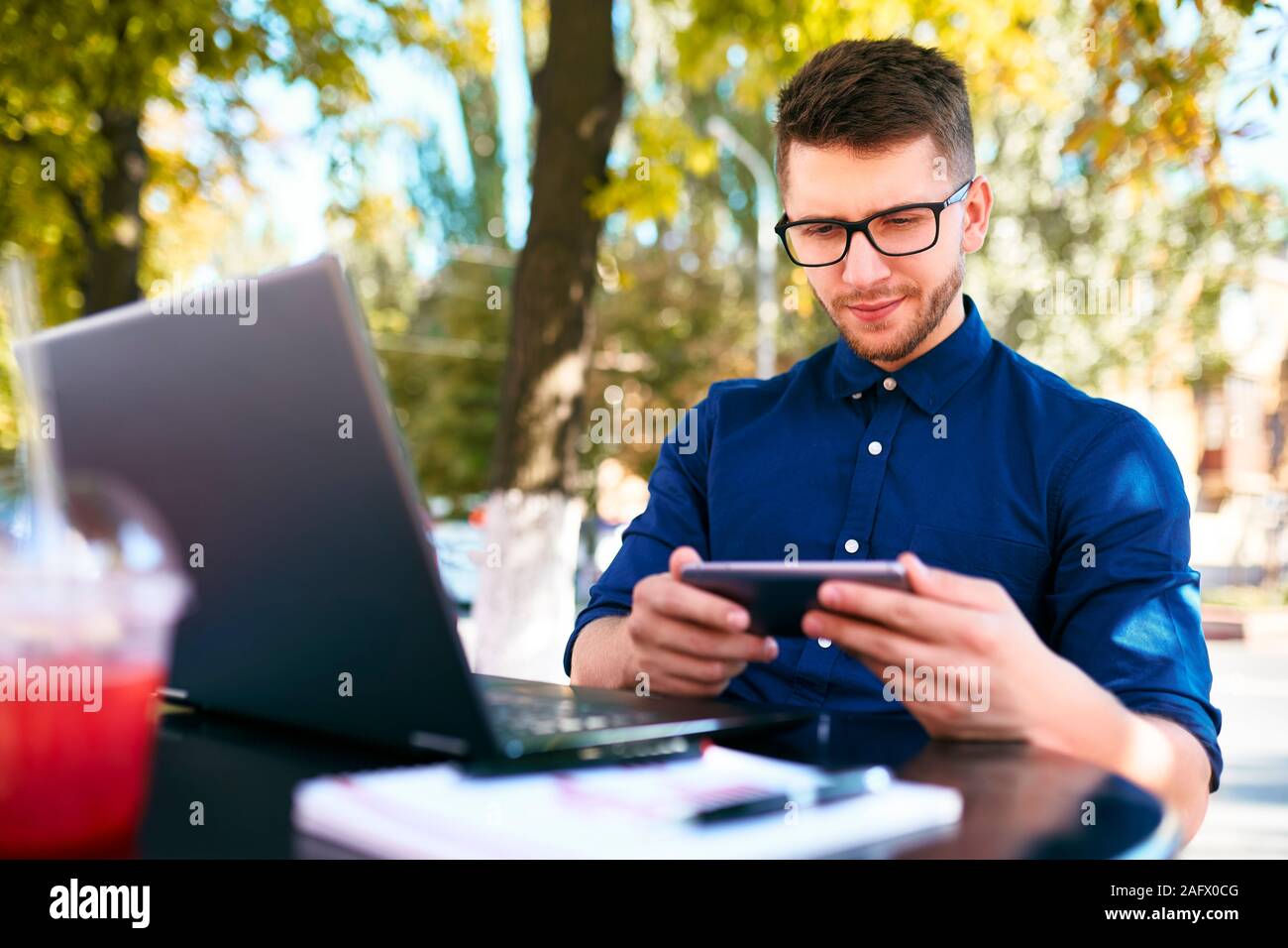 Handsome businessman distracted from work on the laptop watching video on smartphone. Freelancer holding mobile phone and browsing using high speed 4g Stock Photo