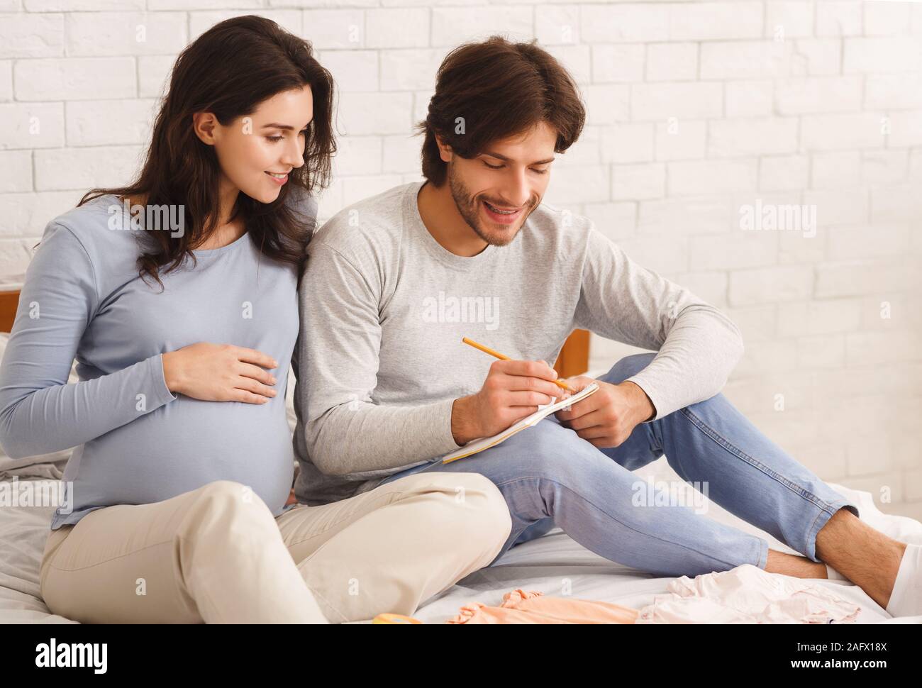 Young pregnant couple choosing baby name while sitting on bed together Stock Photo