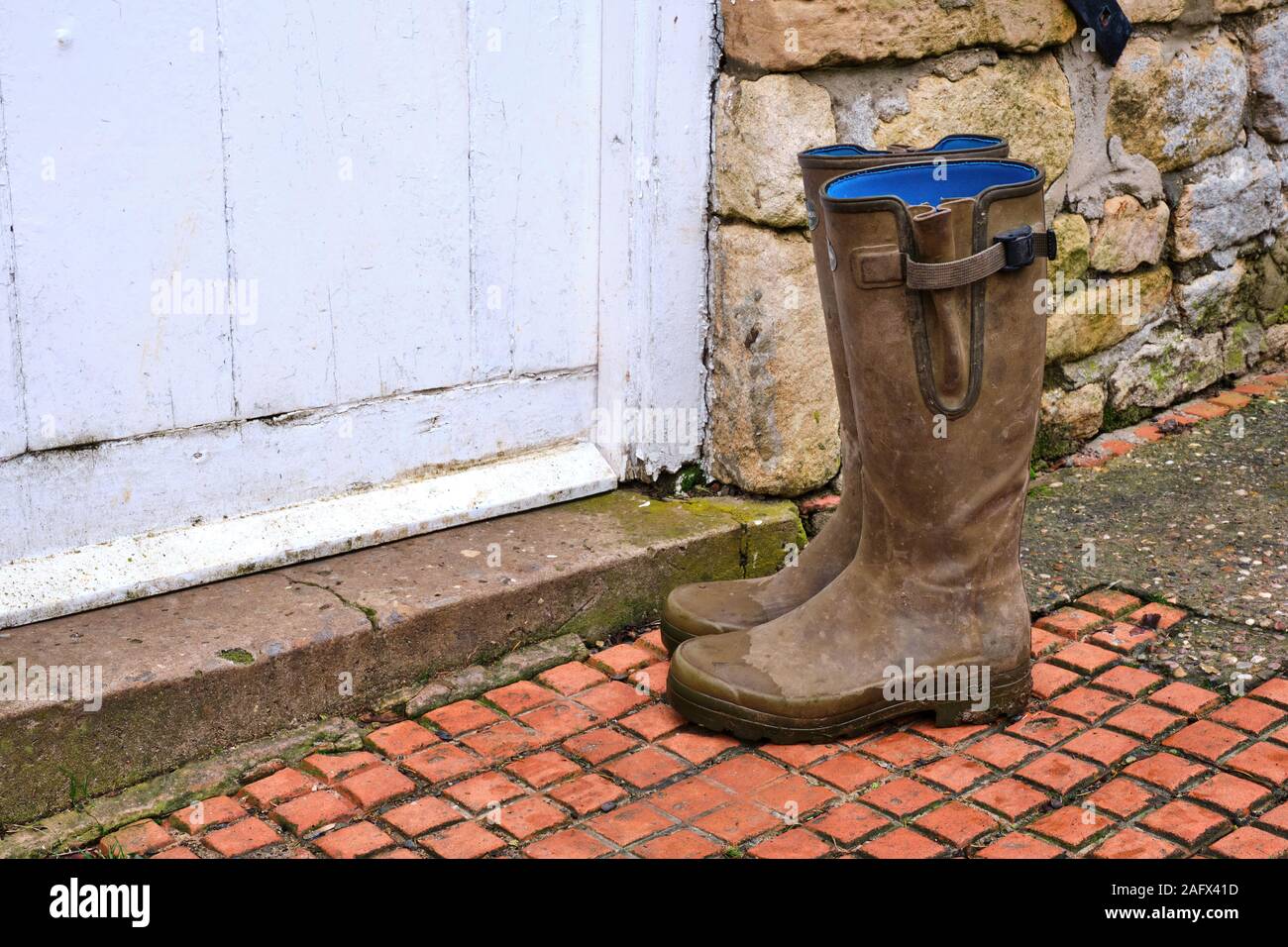 Muddy green wellington boots outside the doorstep of a white door to a stone farm house. Gum boots galoshes rubber boots blucher boot Stock Photo