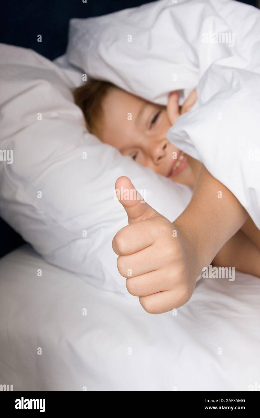 Smiling boy lies on bed with white bed linen and shows thumbs up Stock Photo