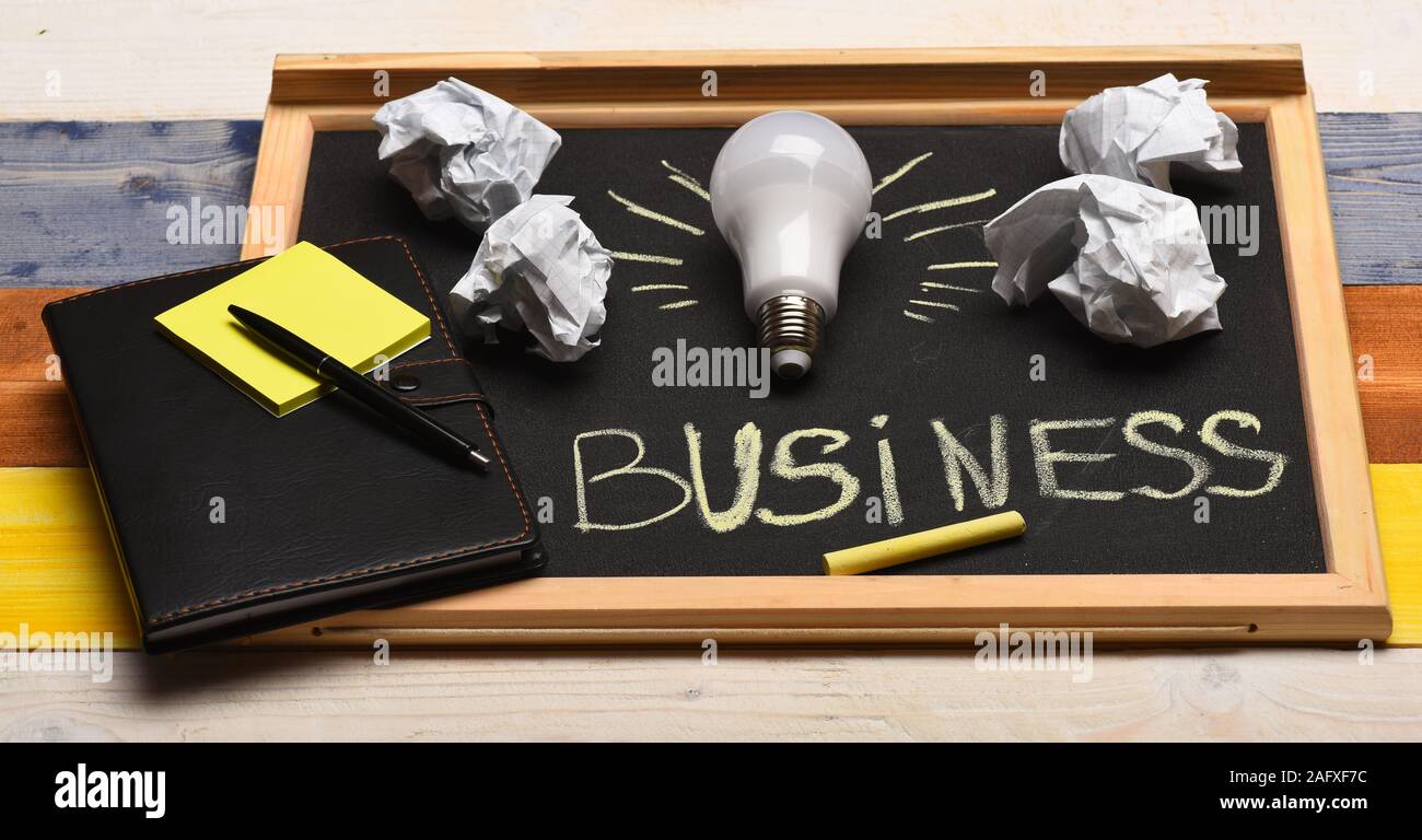 Process of creation: lamp, crumpled paper, chalk and notebook. Board with light bulb, yellow chalk, pen and stickers. Chalkboard with business accessories on wooden background. Business idea concept Stock Photo