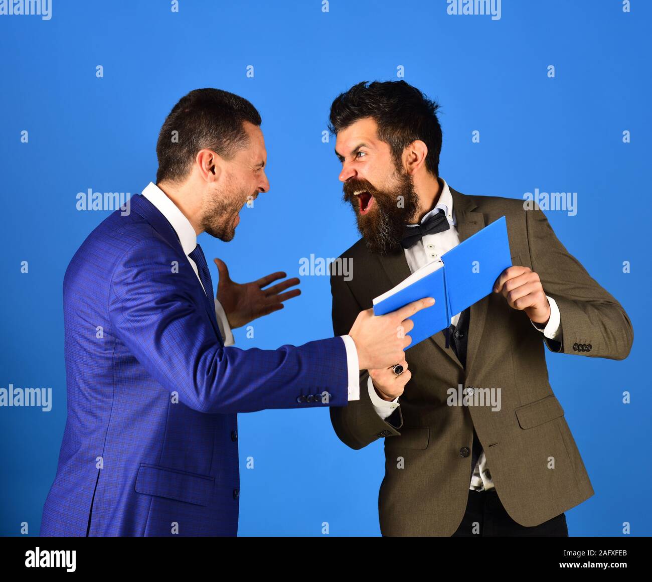 Business conflict and management concept. Machos in classic suits argue about schedule. Men with raging hold blue book. Businessmen with beards yell at each holding business notes Stock Photo -
