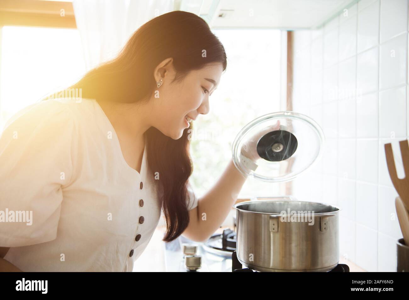 Asian girl teen maid happy cooking soup look inside hot pot at stove in the kitchen in the morning. Stock Photo