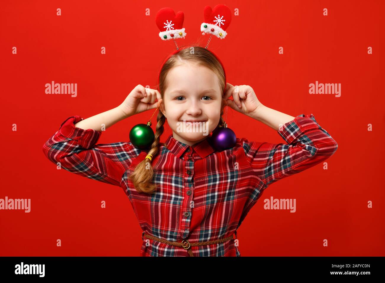 Funny happy little girl with earring christmas ball decoration. A child in a red dress and mittens headband on a colored background. Stock Photo