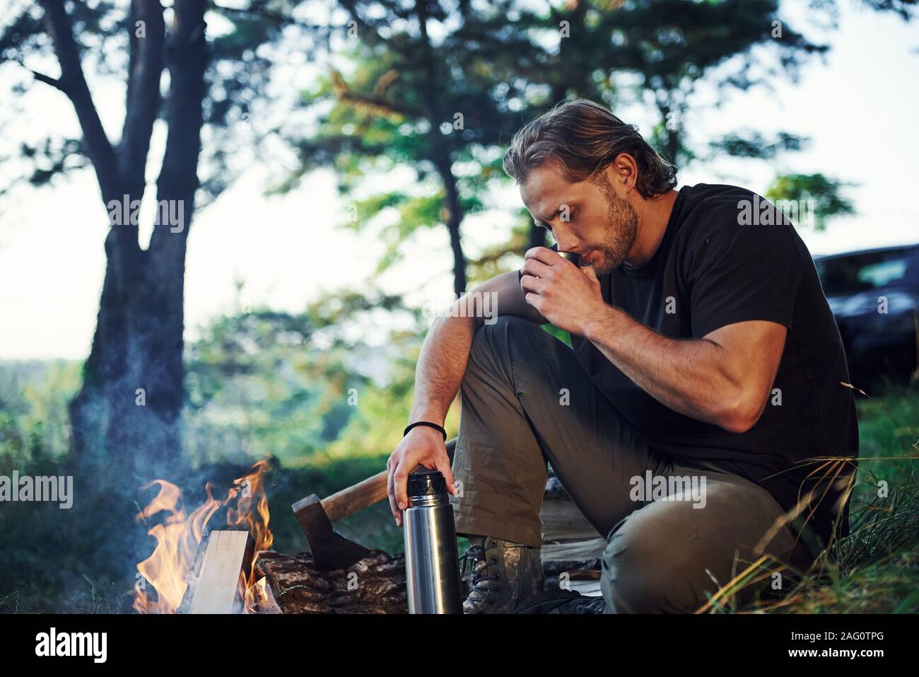 Making drink by using thermos. Man in black shirt near the campfire in the forest at his weekend time Stock Photo