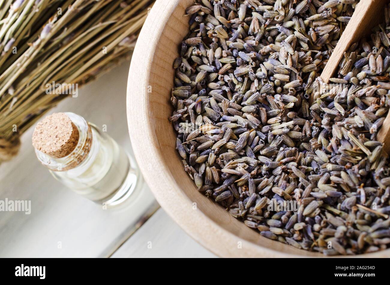 Dried Lavender herbs in wooden bowl with scoop next to essential oil bottle with cork and bunch of flower stems in background. Stock Photo