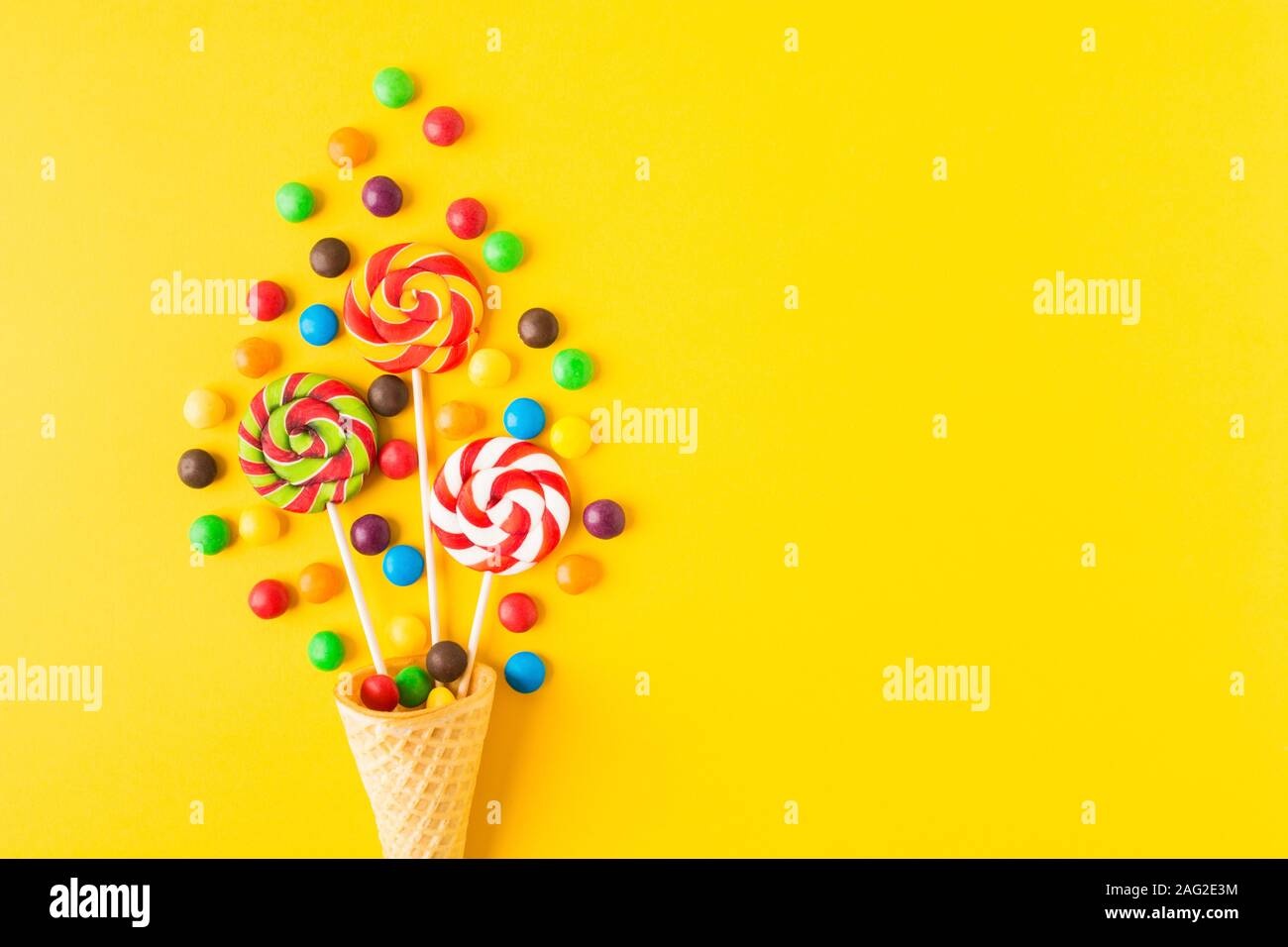 Three colorful round candies on sticks in waffle cone over yellow background with copy space. Top view. Flat lay Stock Photo