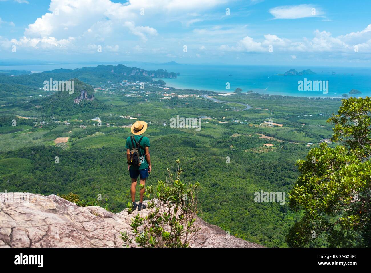 Traveler in hat stands on rock high in mountains landscape & sea on horizon. View of Ao Nang bay and islands from Dragon Crest at Khao Ngon Nak Trail Stock Photo