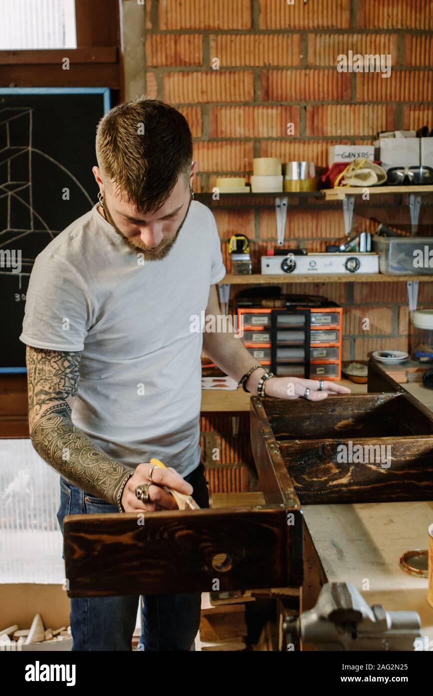 Tattooed artist applying varnish paint on a wooden furniture at craft workshop Stock Photo