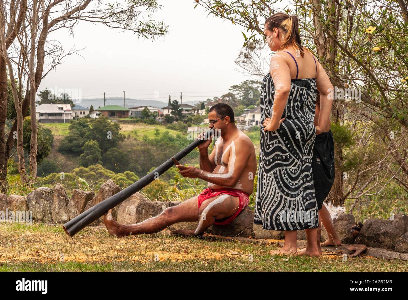 Newcastle, Australia - December 10, 2009: Aboriginal Woman looks at man who sits on tree stump blowing black didgeridoo in front of green foliage. Whi Stock Photo
