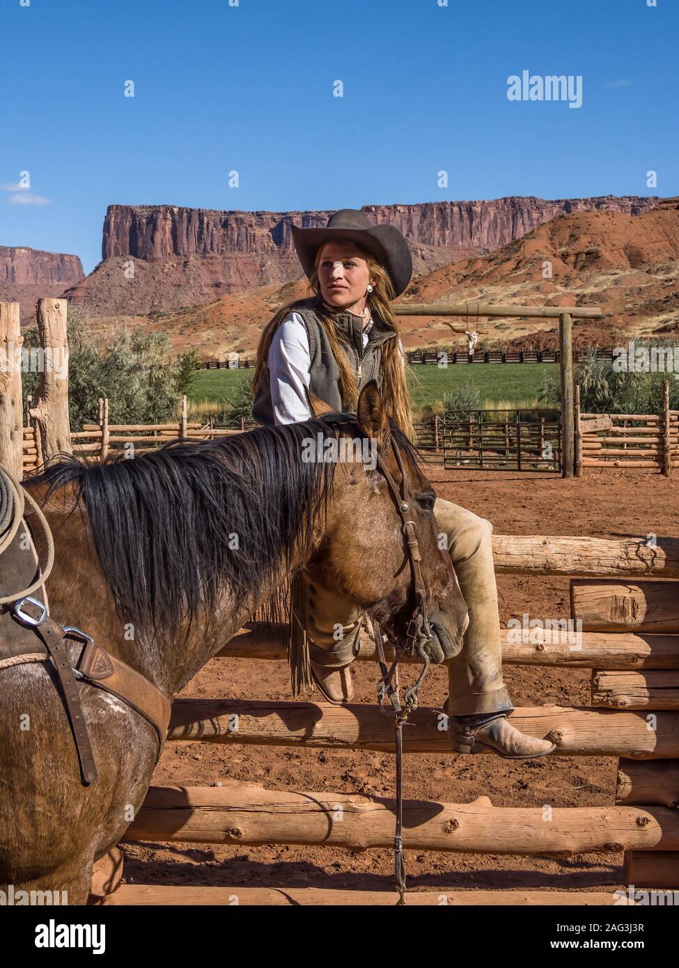 A young attractive working cowgirl wrangler sits on a wood rail fence by her horse on a ranch near Moab, Utah. Stock Photo