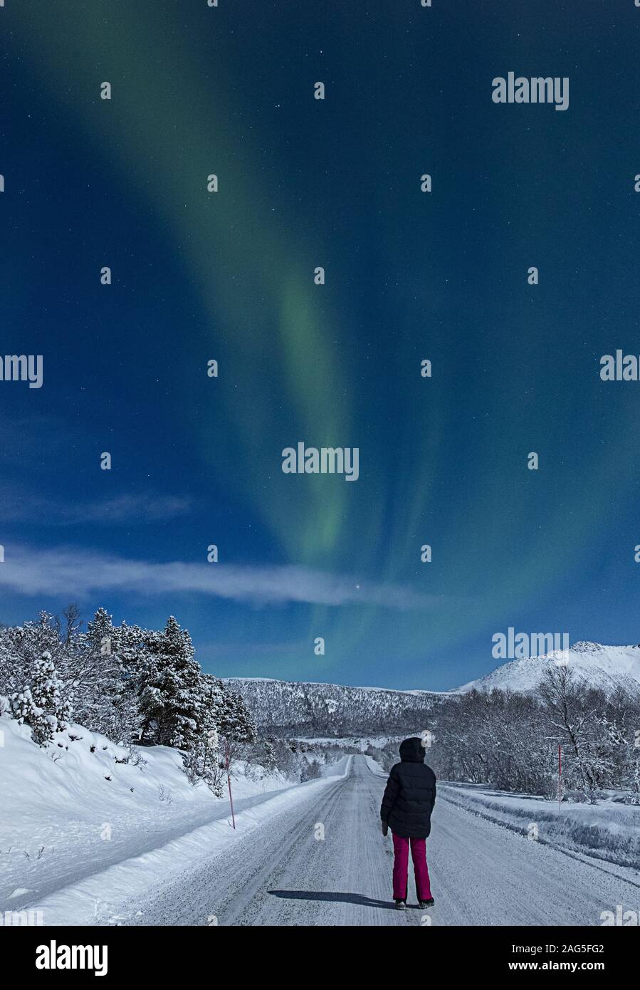 Vertical shot of a person standing on a snow covered road under the northern lights in the sky Stock Photo