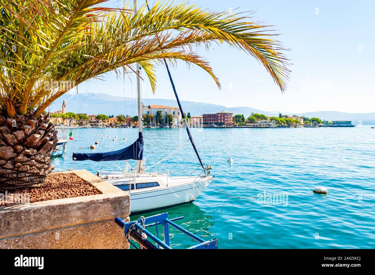 Garda lake western shore, Lombardy Italy. View on Toscolano Maderno cityscape through palm leaves growing on the beach with parked white sailing yacht Stock Photo