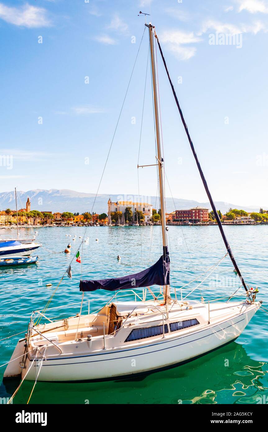 Garda lake western shore, Lombardy Italy. View on parked white sailing yacht on crystal clear blue water of amazing lake Garda and Toscolano Maderno c Stock Photo