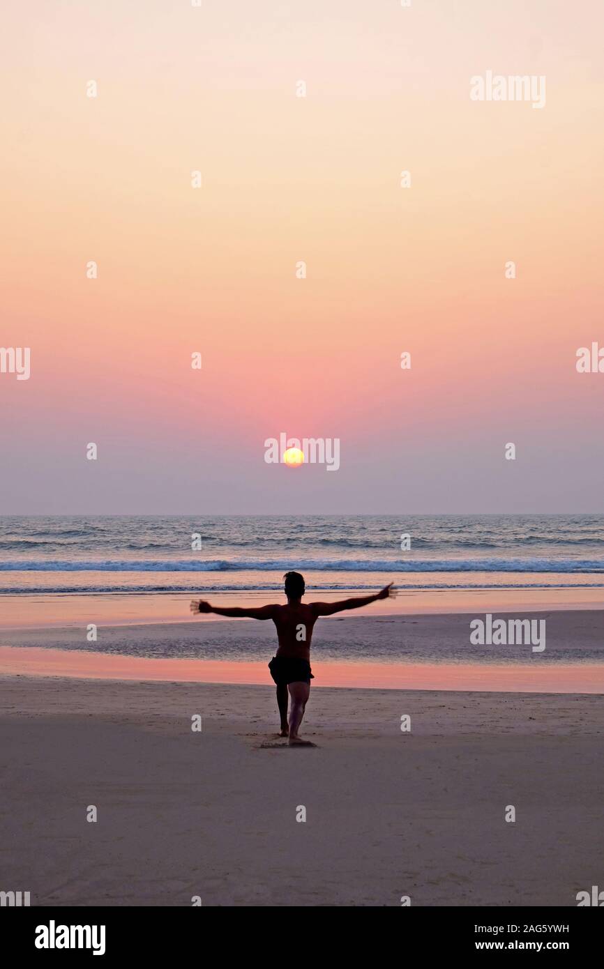 An unrecognizable man doing a yoga pose, the sun salutation on a wide open empty sandy beach, he is kneeling with his arms outstretched, he is lined u Stock Photo