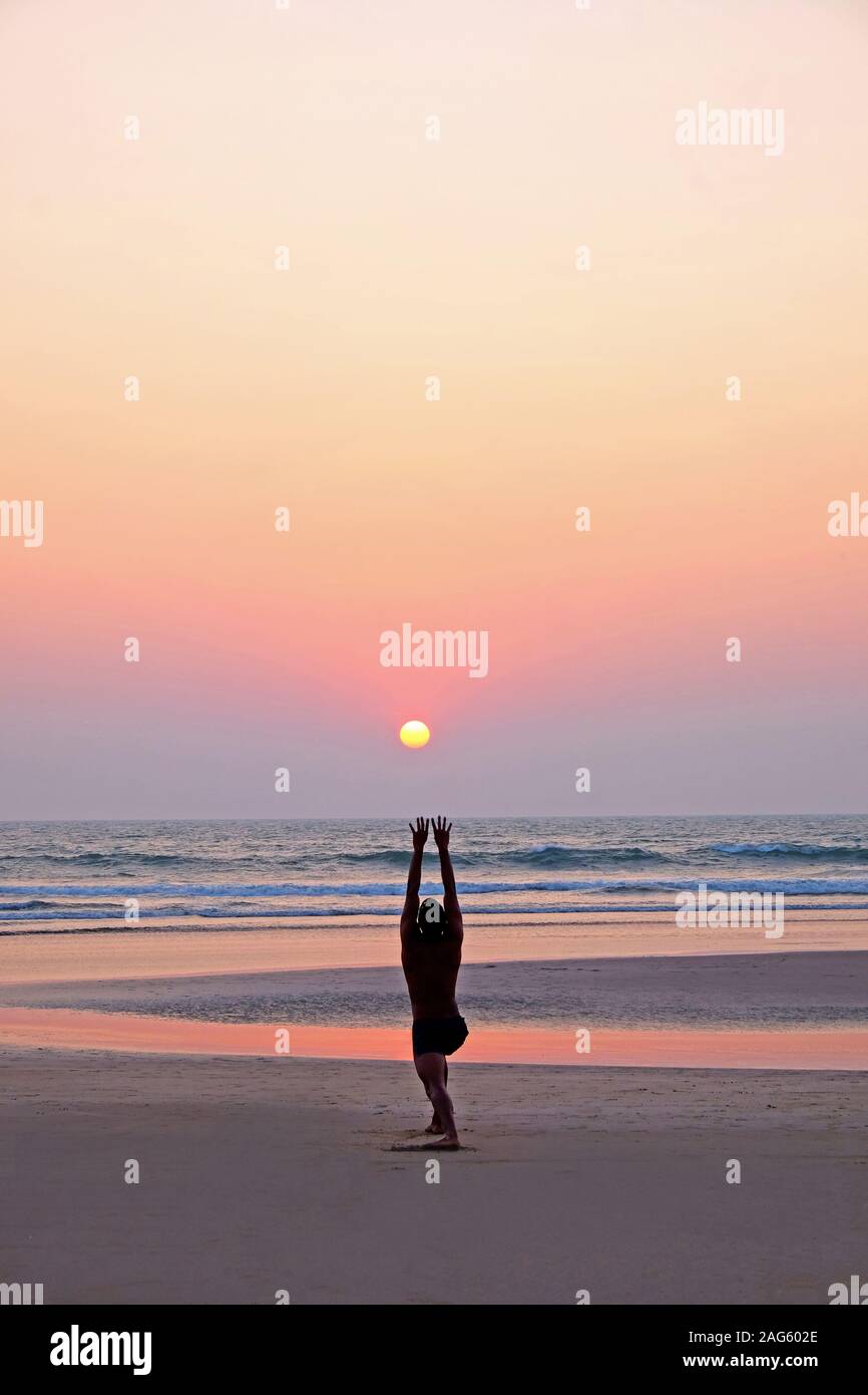 An unrecognizable man doing a yoga pose, the sun salutation, on a wide open empty sandy beach, his legs are bent with his arms outstretched, he is lin Stock Photo