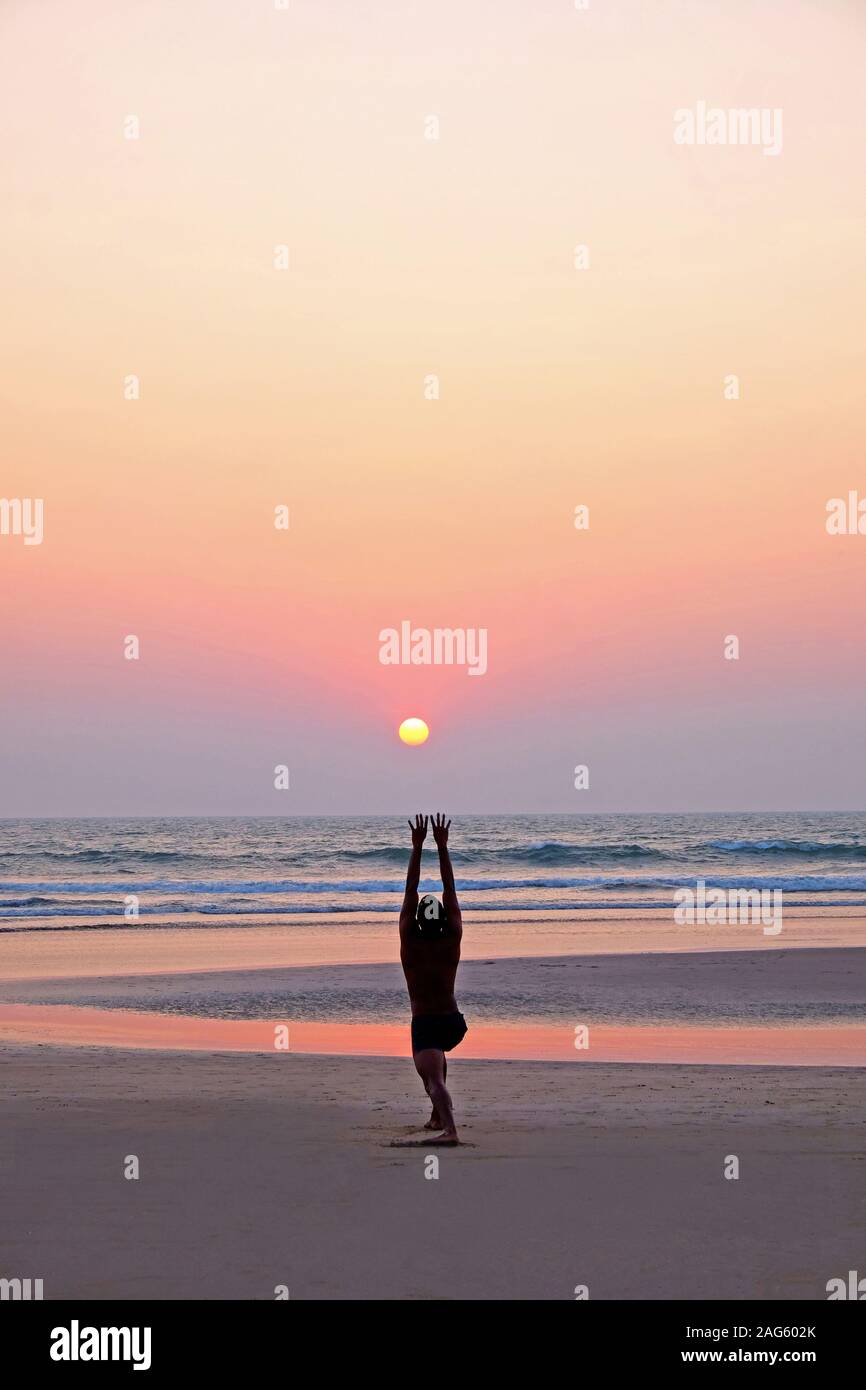 An unrecognizable man doing a yoga pose, the sun salutation on a wide open empty sandy beach, he is bending with his arms stretched up to the sun, the Stock Photo