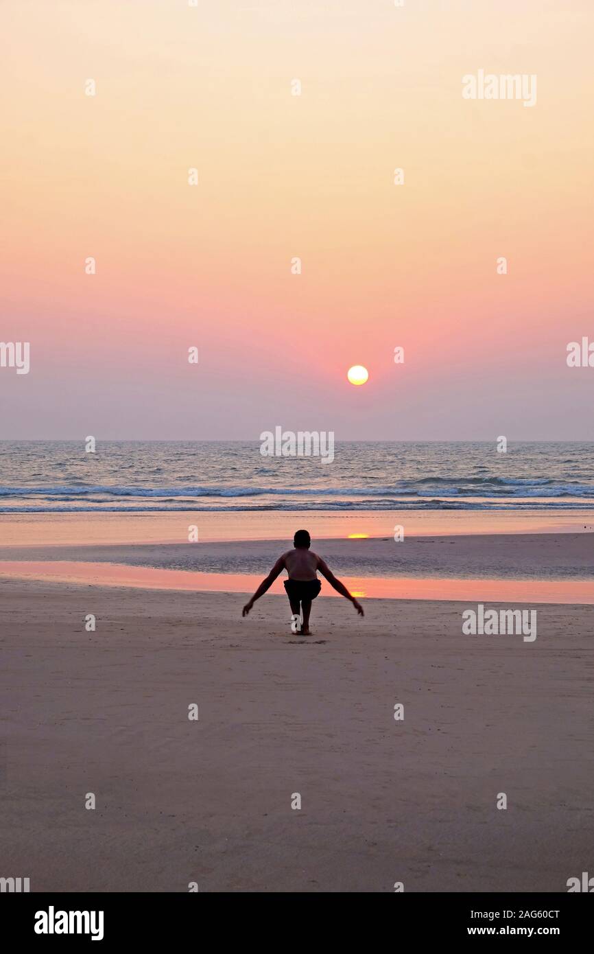 An unrecognizable man doing a yoga pose, the sun salutation on a wide open empty sandy beach, he is bending with his arms outstretched, he is lined up Stock Photo