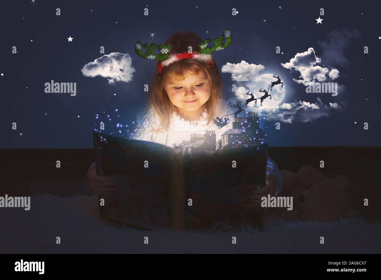 Little girl is reading magic book; Santa sleigh flying over roofs and towards night sky; Christmas magic and tales, background with copy space Stock Photo