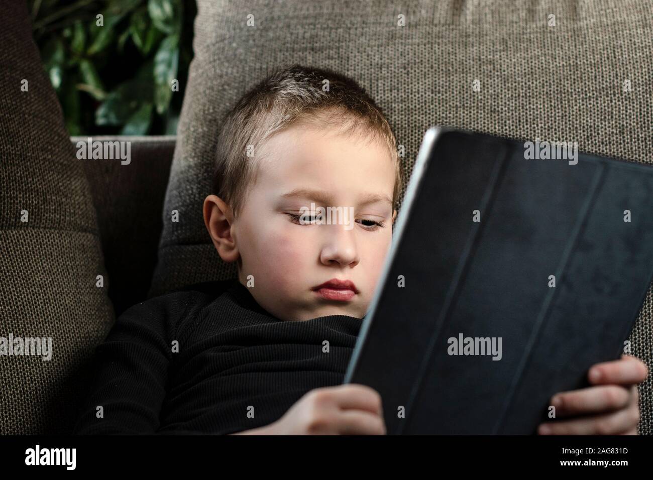 Young boy sitting on sofa in living room and watching cartoons on tablet. Portrait of a smart pre-school child using devices at home. Modern kid. Stock Photo