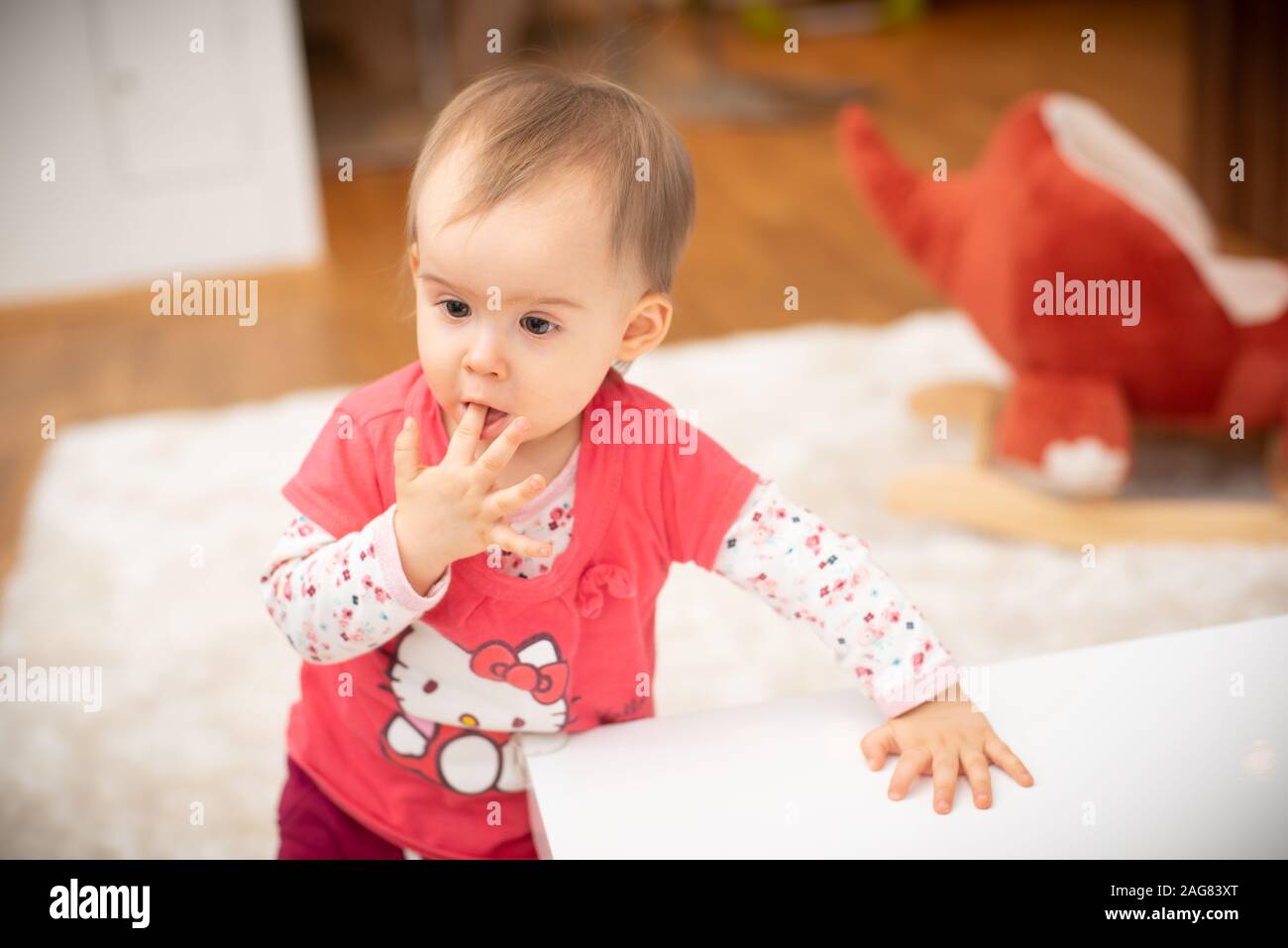Cute 1 year old baby girl bites fingers, concept of teething. Girl in hello kitty red shirt standing Stock Photo