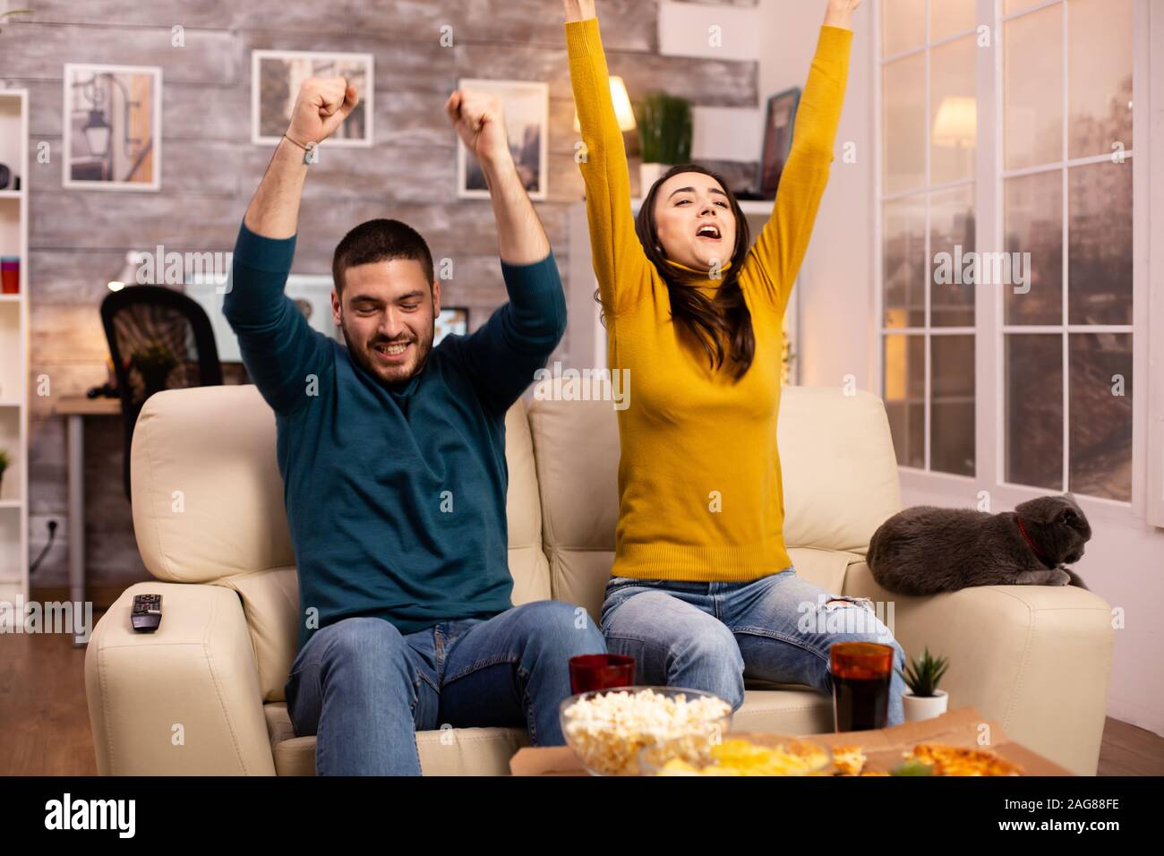 Happy couple cheering for their favourite team while watching TV in he living room. Stock Photo