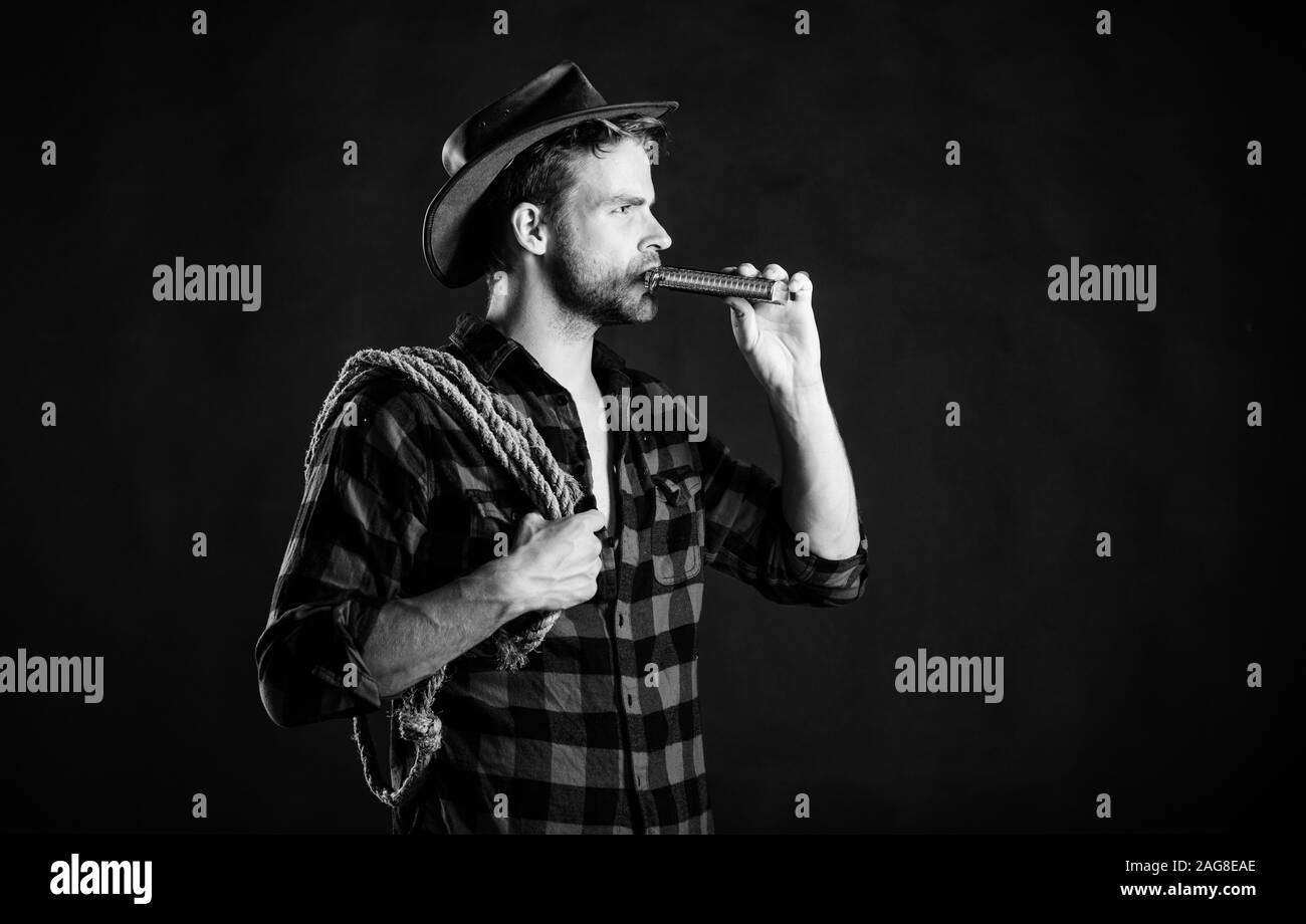 Sheriff concept. Brutal cowboy drinking alcohol. Western culture. Man handsome unshaven cowboy black background. Western life. Man wearing hat hold rope and flask. Lasso tool of American cowboy. Stock Photo