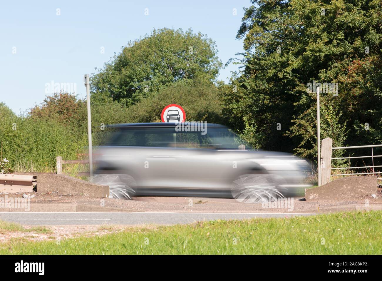 Crick, Northamptonshire, UK: A silver car, blurred by speed, passes a circular No Cars roadsign which is visible just above the car's roof. Stock Photo
