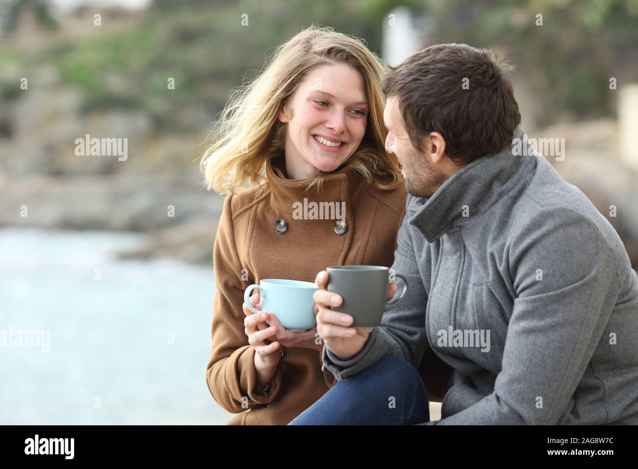 Happy couple of adults talking drinking coffee sitting outdoors in winter on the beach Stock Photo
