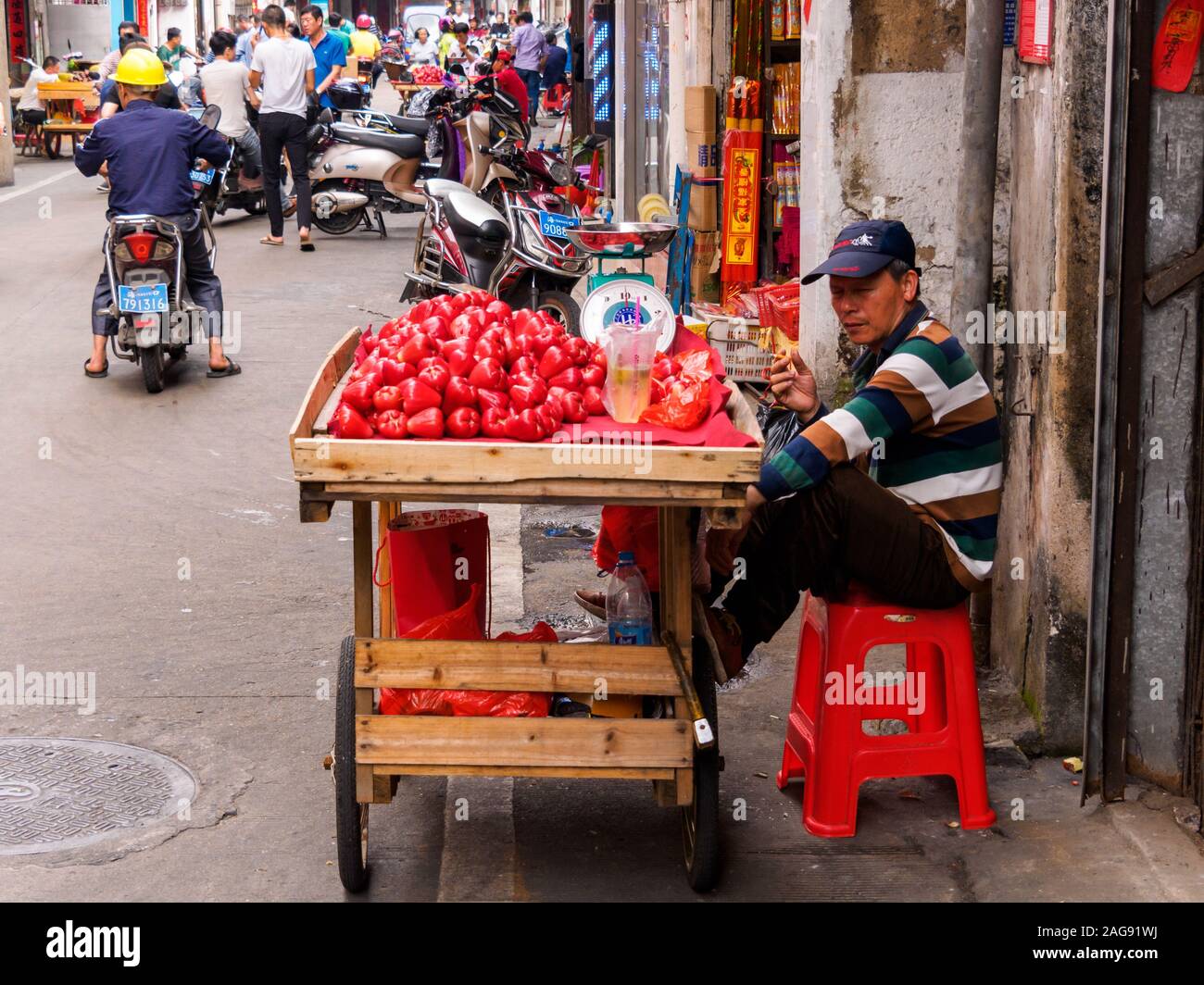 HAIKOU, HAINAN, CHINA - MAR 2 2019 - Street vendor smokes a cigarette while selling tropical wax apples / Jambu fruit from a pushcart Stock Photo
