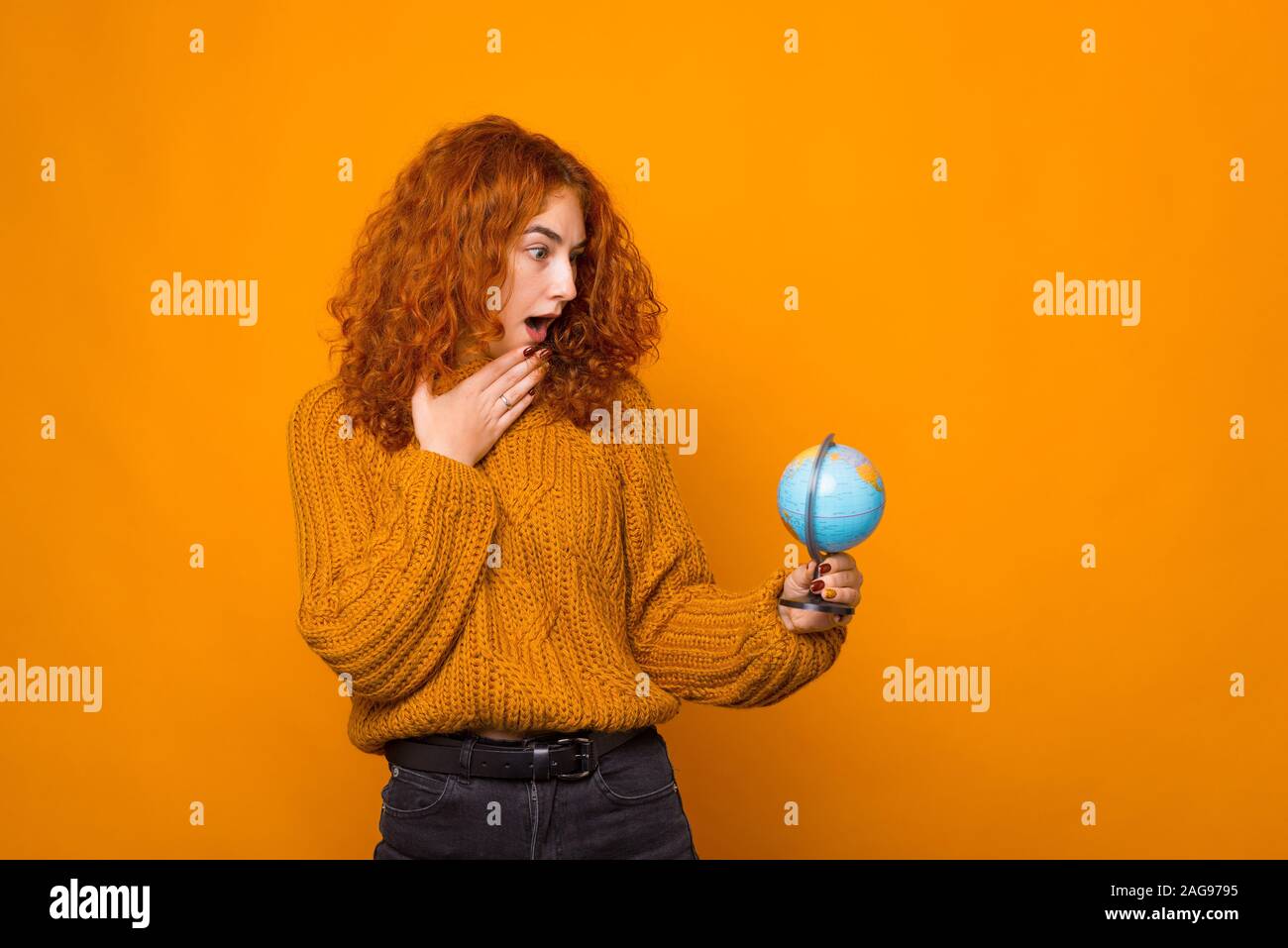 Young woman is looking surprised at globe on orange wall. Stock Photo