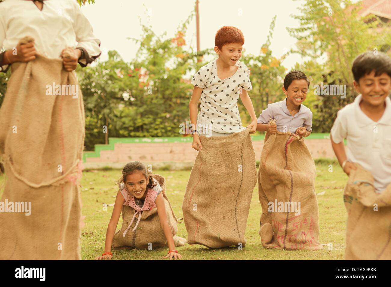 Children's playing potato sack jumping race at park outdoor - kids falling and having fun while playing gunny sack race. Stock Photo