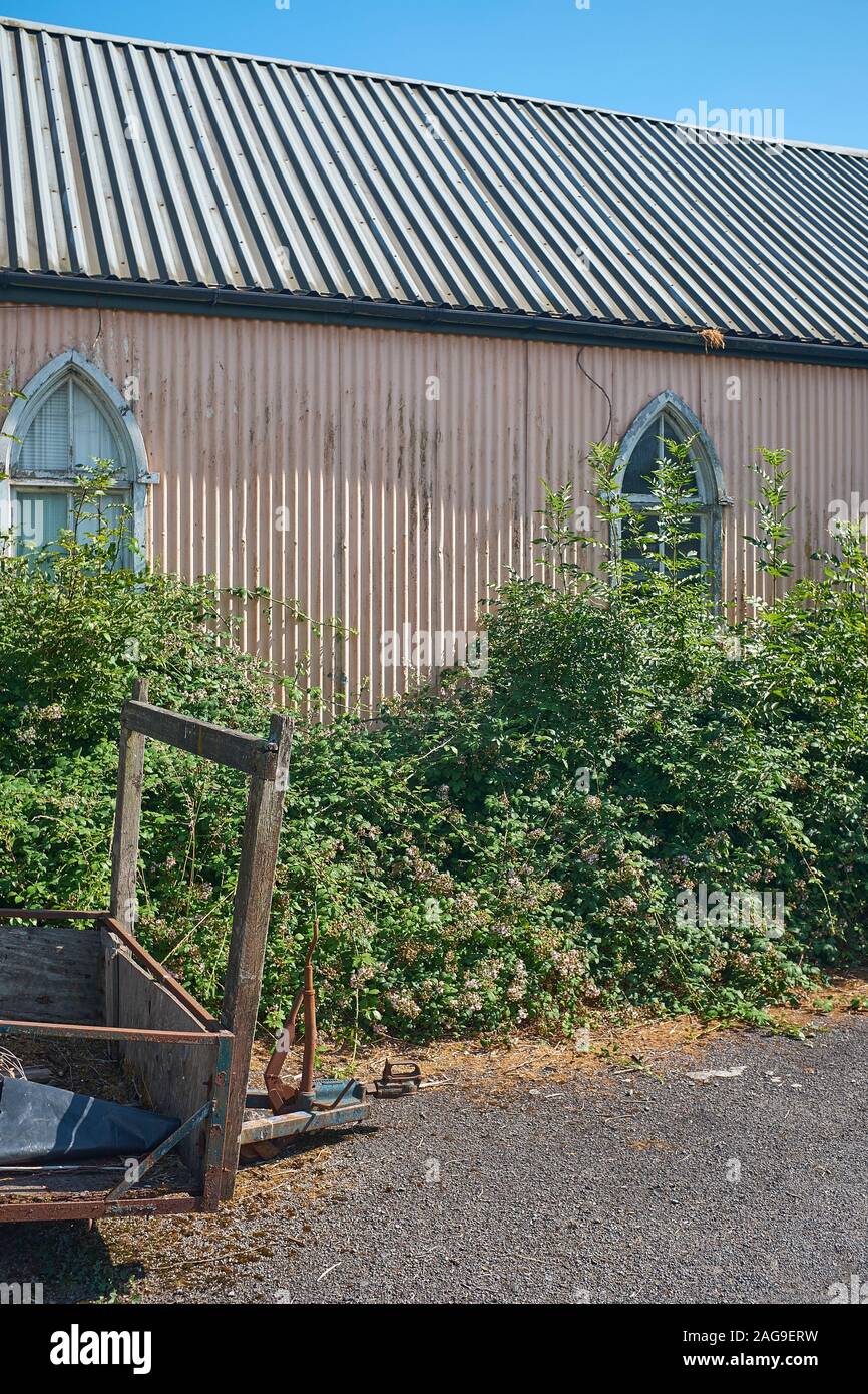 An old building that has been made from corrugated sheeting that has arched windows, an overgrown hedge and an old worn out trailer outside Stock Photo