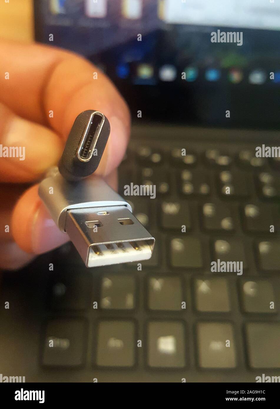 Male hand holds a usb-c and usb 3.0 connection above a notebook computer with screen on and with a black keyboard. Stock Photo