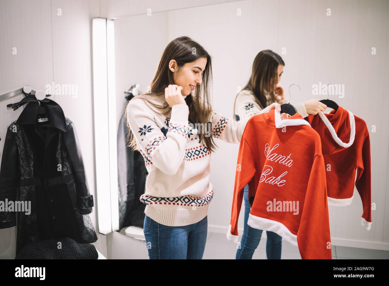 Slim girl looking new red sweater on hanger. Smiling girl holding Santa baby sweater on hanger in dressing room with mirror, with copyspace. Image can Stock Photo