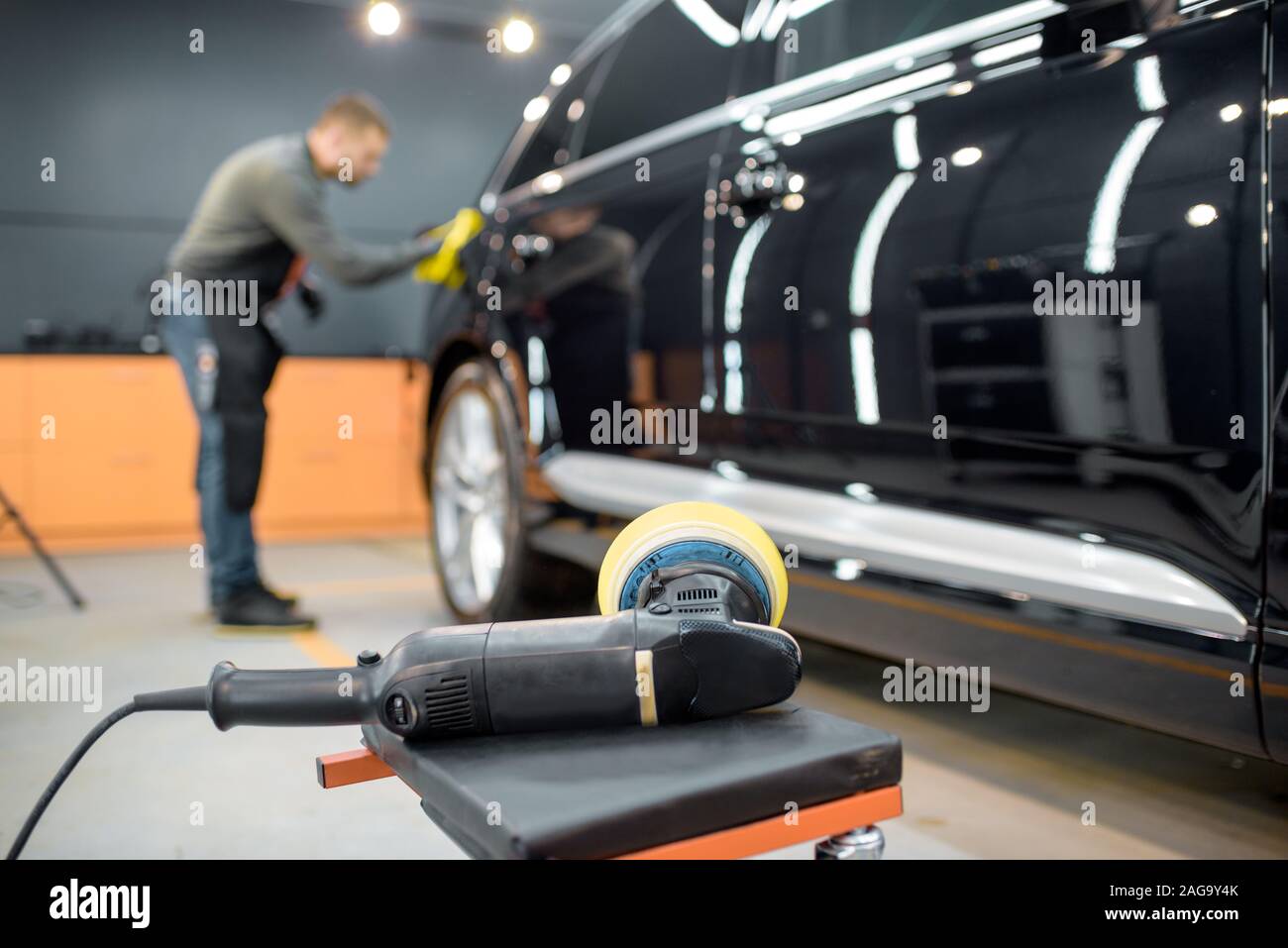 Car service worker wiping vehicle body with microfiber, examining glossy coating after the polishing procedure. Grinder on the foreground Stock Photo